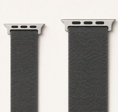 Zwei Apple-Watch-Armbänder aus anthrazitfarbenem Nylon nebeneinander auf weißem Grund: links ein schmales Band der kleinen Größengruppe, rechts ein breiteres der großen Gruppe, jeweils mit dem flachen Anschlussstück. Mit KI erstellte Materialdarstellung.