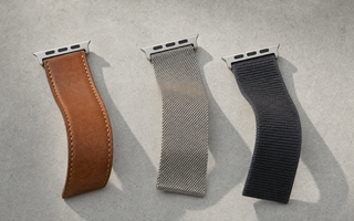 Drei Apple-Watch-ArmbĂ€nder in leichten S-Kurven auf hellem Beton: cognacbraunes Leder, Edelstahl-Milanaise und anthrazitfarbenes Nylon, jeweils mit dem flachen AnschlussstĂŒck. Mit KI erstellte Materialdarstellung.
