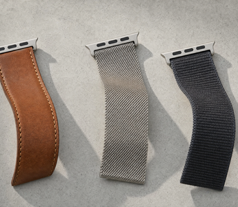 Drei Apple-Watch-Armbänder in leichten S-Kurven auf hellem Beton: cognacbraunes Leder, Edelstahl-Milanaise und anthrazitfarbenes Nylon, jeweils mit dem flachen Anschlussstück. Mit KI erstellte Materialdarstellung.