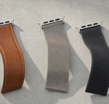 Drei Apple-Watch-Armbänder in leichten S-Kurven auf hellem Beton: cognacbraunes Leder, Edelstahl-Milanaise und anthrazitfarbenes Nylon, jeweils mit dem flachen Anschlussstück. Mit KI erstellte Materialdarstellung.
