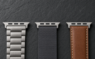 Drei Apple-Watch-ArmbĂ€nder aus gebĂŒrstetem Titan, anthrazitfarbenem Nylon und cognacbraunem Leder nebeneinander auf dunklem Schiefer, jedes mit dem breiten flachen AnschlussstĂŒck am oberen Bandende. Mit KI erstellte Materialdarstellung.