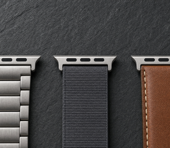 Drei Apple-Watch-Armbänder aus gebürstetem Titan, anthrazitfarbenem Nylon und cognacbraunem Leder nebeneinander auf dunklem Schiefer, jedes mit dem breiten flachen Anschlussstück am oberen Bandende. Mit KI erstellte Materialdarstellung.