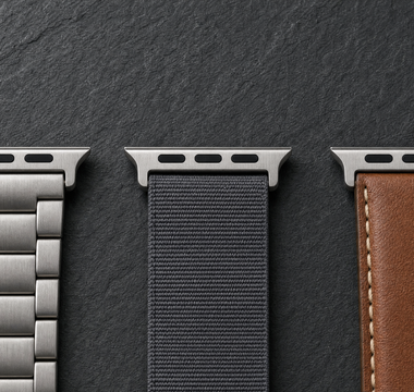 Drei Apple-Watch-Armbänder aus gebürstetem Titan, anthrazitfarbenem Nylon und cognacbraunem Leder nebeneinander auf dunklem Schiefer, jedes mit dem breiten flachen Anschlussstück am oberen Bandende. Mit KI erstellte Materialdarstellung.