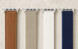 Vier Apple-Watch-ArmbĂ€nder nebeneinander auf hellem Kalkstein: braunes Leder, champagnerfarbenes Milanaise-Geflecht, weiĂes Silikon und dunkelblaues Nylon, jedes mit dem flachen AnschlussstĂŒck. Mit KI erstellte Materialdarstellung.