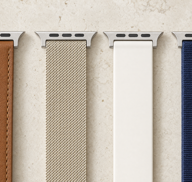 Vier Apple-Watch-Armbänder nebeneinander auf hellem Kalkstein: braunes Leder, champagnerfarbenes Milanaise-Geflecht, weißes Silikon und dunkelblaues Nylon, jedes mit dem flachen Anschlussstück. Mit KI erstellte Materialdarstellung.