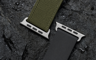 Zwei robuste Apple-Watch-ArmbĂ€nder auf nassem dunklem Basalt: ein olivgrĂŒnes Gewebeband und ein schwarzes geripptes Kautschukband, beide mit dem flachen AnschlussstĂŒck. Mit KI erstellte Materialdarstellung.