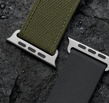 Zwei robuste Apple-Watch-Armbänder auf nassem dunklem Basalt: ein olivgrünes Gewebeband und ein schwarzes geripptes Kautschukband, beide mit dem flachen Anschlussstück. Mit KI erstellte Materialdarstellung.