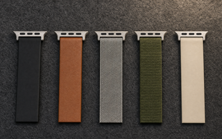 FĂŒnf Armbandabschnitte nebeneinander auf dunklem Filz: schwarzes Silikon, braunes Leder, Edelstahlgeflecht, olivgrĂŒnes Nylon und sandfarbenes Silikon, jeweils mit AnschlussstĂŒck. Mit KI erstellte Materialdarstellung.