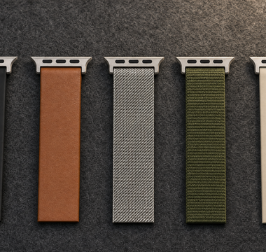 Fünf Armbandabschnitte nebeneinander auf dunklem Filz: schwarzes Silikon, braunes Leder, Edelstahlgeflecht, olivgrünes Nylon und sandfarbenes Silikon, jeweils mit Anschlussstück. Mit KI erstellte Materialdarstellung.