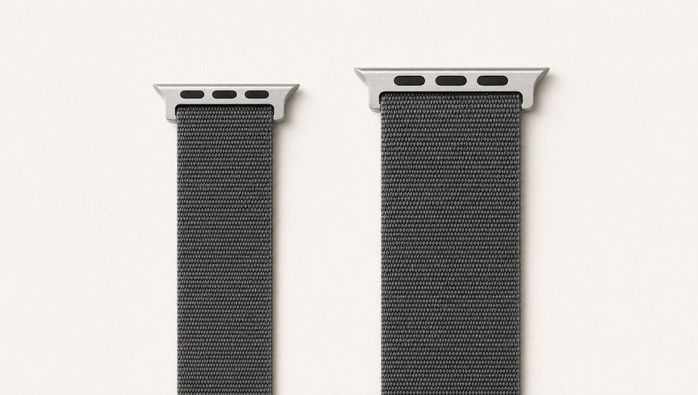 Zwei Apple-Watch-Armbänder aus anthrazitfarbenem Nylon nebeneinander auf weißem Grund: links ein schmales Band der kleinen Größengruppe, rechts ein breiteres der großen Gruppe, jeweils mit dem flachen Anschlussstück. Mit KI erstellte Materialdarstellung.