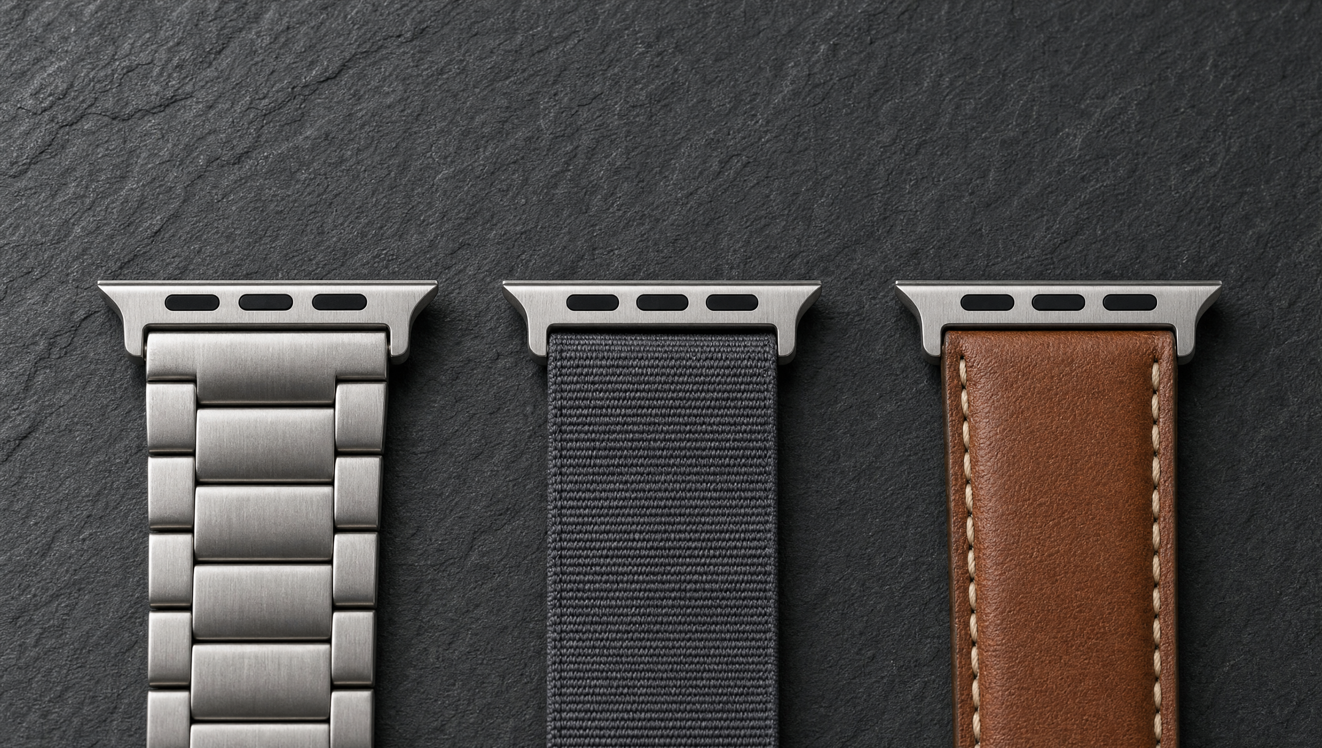 Drei Apple-Watch-Armbänder aus gebürstetem Titan, anthrazitfarbenem Nylon und cognacbraunem Leder nebeneinander auf dunklem Schiefer, jedes mit dem breiten flachen Anschlussstück am oberen Bandende. Mit KI erstellte Materialdarstellung.
