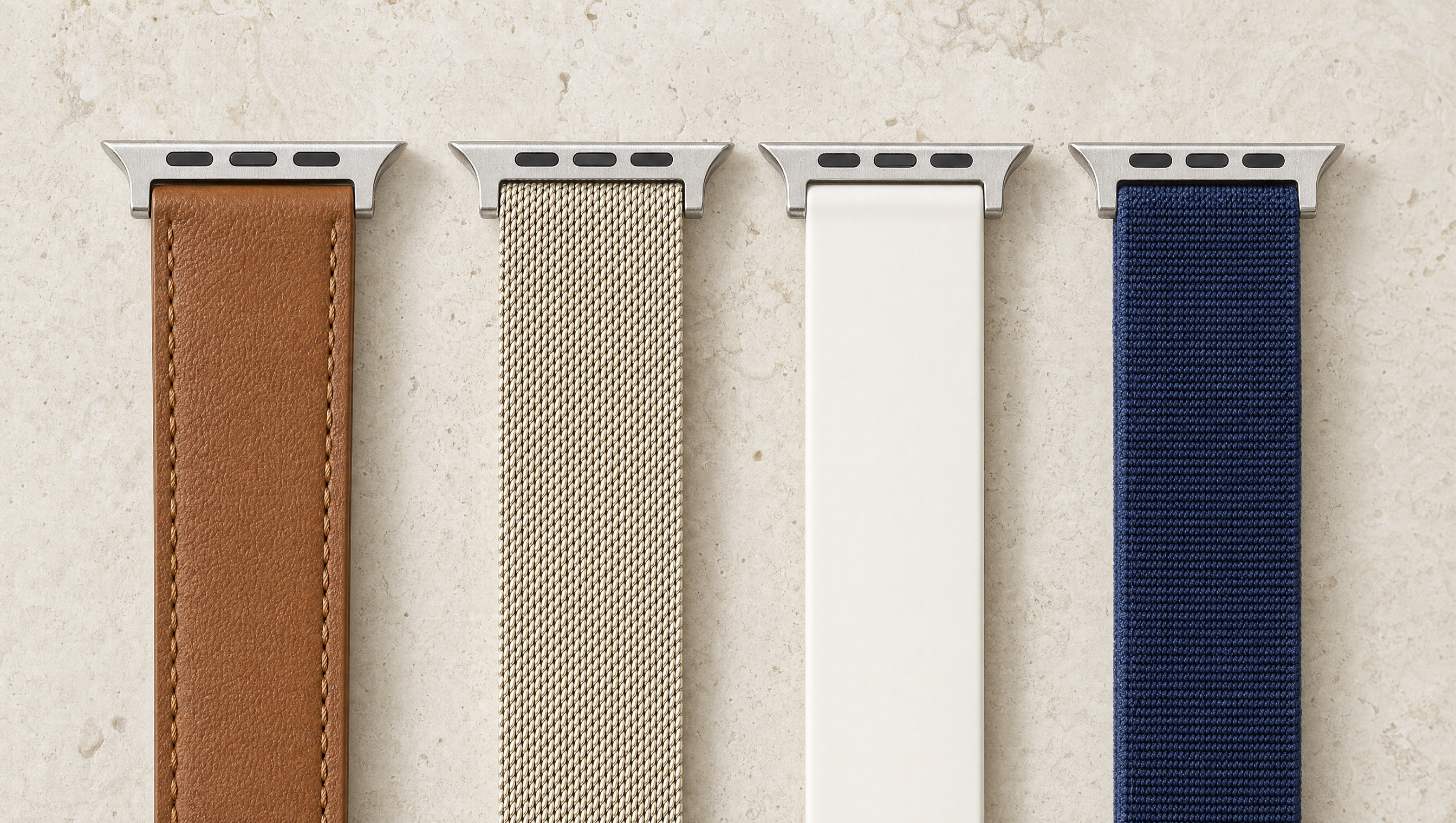 Vier Apple-Watch-Armbänder nebeneinander auf hellem Kalkstein: braunes Leder, champagnerfarbenes Milanaise-Geflecht, weißes Silikon und dunkelblaues Nylon, jedes mit dem flachen Anschlussstück. Mit KI erstellte Materialdarstellung.