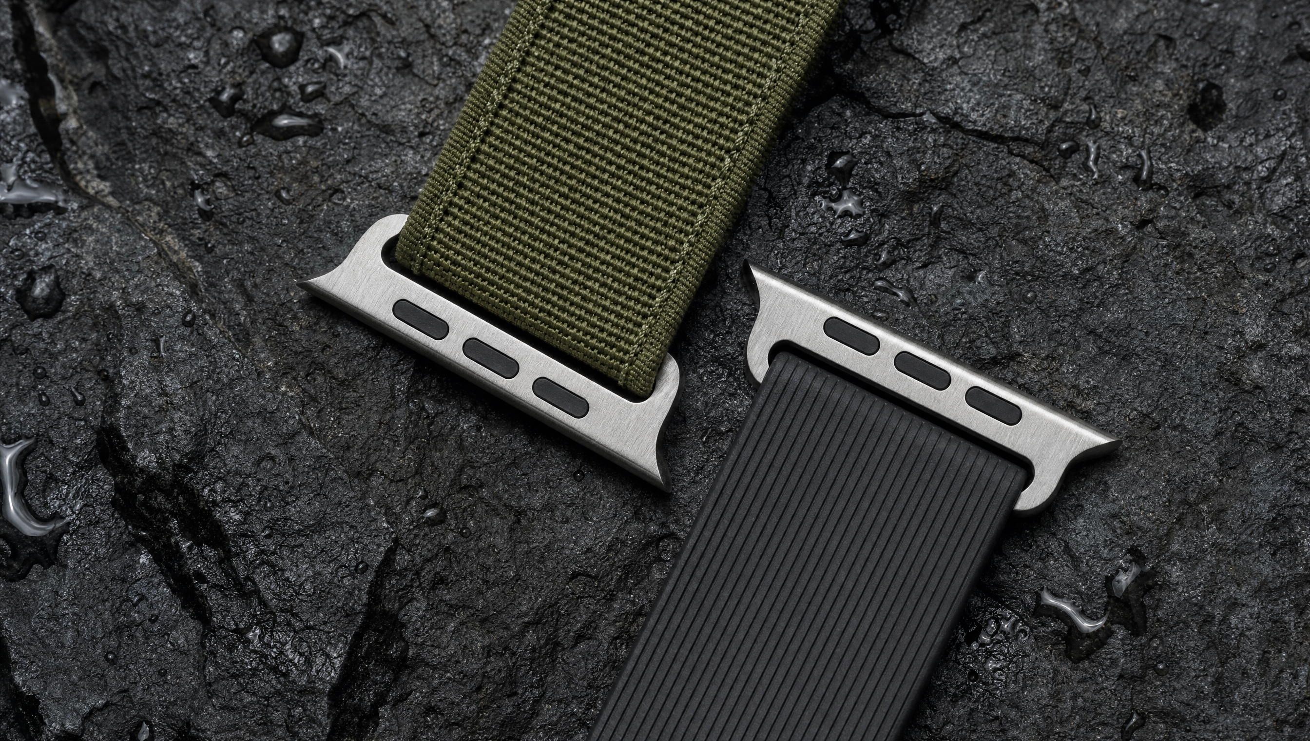 Zwei robuste Apple-Watch-Armbänder auf nassem dunklem Basalt: ein olivgrünes Gewebeband und ein schwarzes geripptes Kautschukband, beide mit dem flachen Anschlussstück. Mit KI erstellte Materialdarstellung.